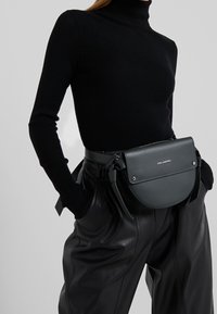 Zwarte geribbelde coltrui gecombineerd met hooggetailleerde zwarte leren broek. Voorzien van een groene crossbodytas met een gebogen ontwerp en ritssluiting.