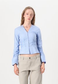 Blusa azzurro chiaro a maniche lunghe con scollo a V, texture arricciata e maniche a campana. Abbinata a pantaloni beige. Tessuto liscio e lavorato.