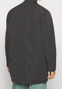 Cappotto lungo nero con colletto alto, texture liscia e dettagli di cucitura visibili. Presenta una vestibilità ampia e una leggera lucentezza.