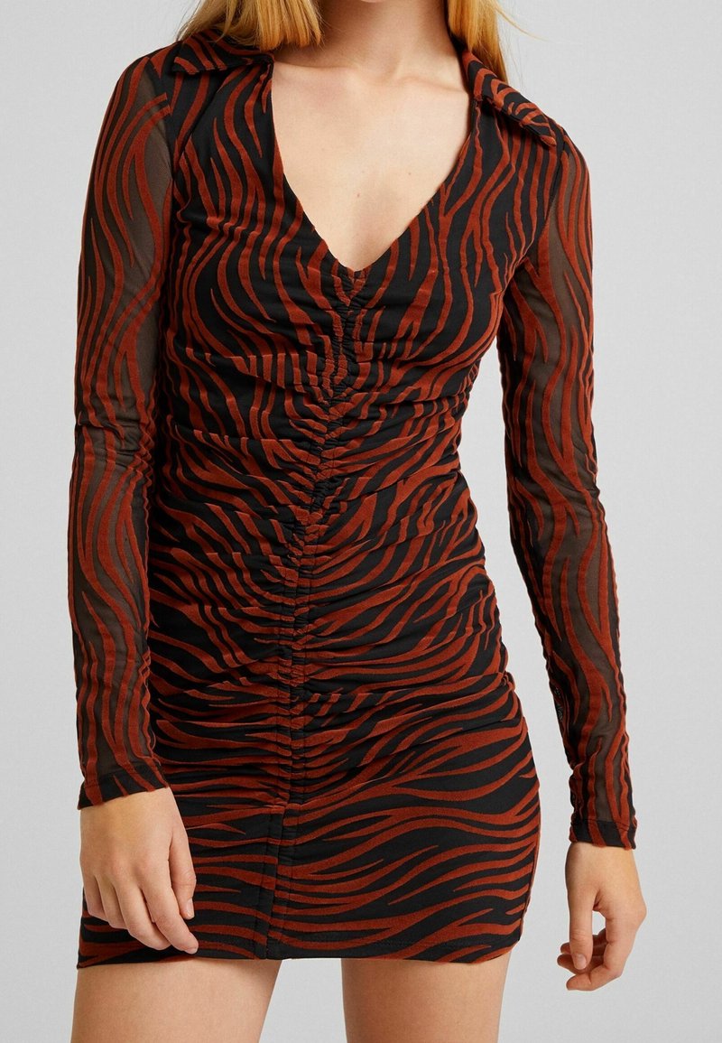 Kobieta nosząca mini sukienkę z długim rękawem w czarno-rustową zebrę, z głębokim dekoltem w kształcie litery V, na jednolitym tle.