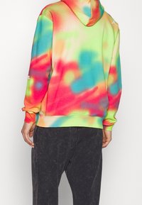 Personne portant un sweat à capuche tie-dye aux couleurs vives avec une capuche et un pantalon décontracté à lavage foncé, vue de dos.