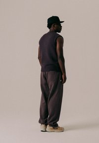 Ärmelloser lila strukturierter Strickpullover, lässige dunkelgraue Hose und beige Sneaker, von hinten betrachtet. Schwarze Mütze und Sonnenbrille getragen.
