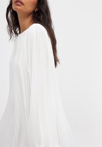 Valkoinen laskostettu mekko pitkillä hihoilla, jossa on pyöreä pääntie ja pehmeä, virtaava rakenne, joka esittelee pystysuoria viivoja kankaassa.