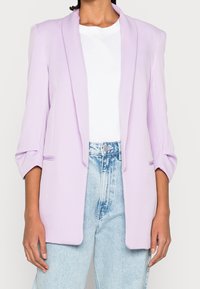 Blazer lilas avec col châle, manches longues et deux poches latérales, associé à un t-shirt blanc et un jean taille haute bleu clair.