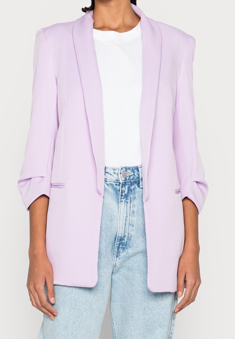 Blazer lilas avec col châle, manches longues et deux poches latérales, associé à un t-shirt blanc et un jean taille haute bleu clair.