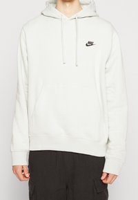 Ljusgrå Nike-hoodie i mjuk bomullsblandning. Den har en framficka med dragkedja, justerbara dragsnören och en liten svart logotyp på bröstet.
