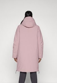 Lange roze gewatteerde jas met een capuchon, voorzien van een soepele textuur en zijritsdetails, gedragen over een grijze broek.