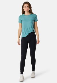T-shirt de sport teal à manches courtes, associé à un legging noir avec poches latérales. Modèle portant des baskets blanches, se tenant debout.