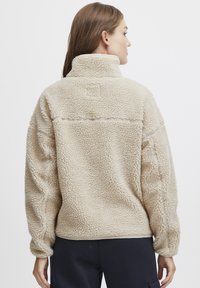 Beige fleece genser med høy krage, teksturert, løs passform, med en horisontal søm over den øvre ryggen og elastiske mansjetter.