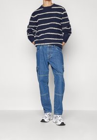 Marinblå randig stickad tröja med vita linjer, ihop med blå cargojackor med sidofickor och kontrasterande sömmar, bärs med vita sneakers.