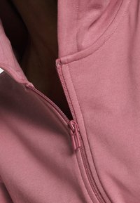 Haut à zip rose avec une texture douce et côtelée, avec une fermeture éclair et un col assortis, affiché partiellement avec le zip ouvert.