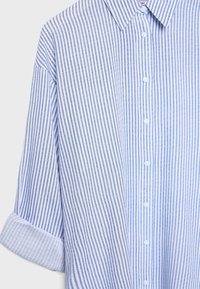 Camicia a righe verticali blu e bianche con bottoni, colletto e maniche arrotolate su uno sfondo chiaro.