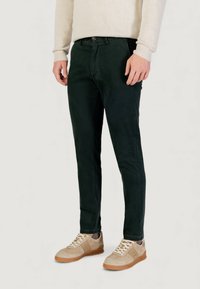 Pantalones verde oscuro de algodón, con un diseño ajustado, bolsillos delanteros y cierre con botón. Combinados con zapatillas beige.
