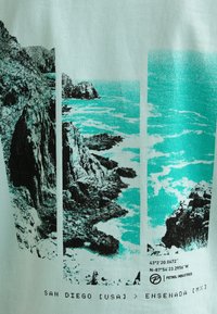 T-shirt verde menta con una grafica nera e verde teal che rappresenta coste rocciose e onde oceaniche, con testo che dettaglia luoghi e coordinate.