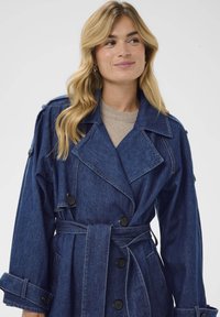 Denim plašč v temno modri barvi, z dvojnim gumbom na sprednji strani, širokim ovratnikom in pasom. Vsebuje dve stranski žepi.