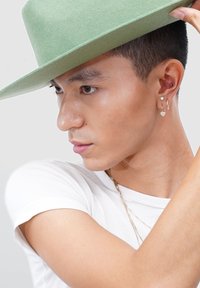 Young man in white shirt adjusting light green wide-brimmed hat, wearing gold hoop earrings, a stud, and delicate layered necklaces.