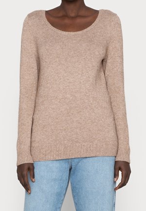 Osoba o ciemnej karnacji, ubrana w beżowy, dziergany sweter z dekoltem w serek oraz jasnoniebieskie dżinsy, stojąca z ramionami swobodnie opuszczonymi wzdłuż ciała.