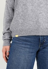Pull en laine gris avec des poignets et un ourlet côtelés, associé à un jean en denim bleu. Petite étiquette jaune au bas du pull. Texture lisse.
