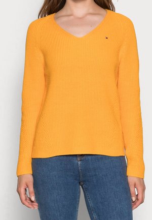 Femme portant un pull jaune à manches longues et col en V et un jean bleu, debout, les bras détendus le long du corps.