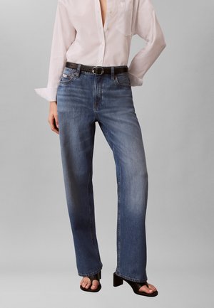 Persona che indossa jeans dritti blu scoloriti, camicia bianca con bottoni, cintura nera e sandali neri con tacco e punta aperta su sfondo semplice.