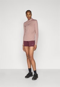 Gaiši rozā sporta krekls ar garām piedurknēm un ceturkšņa rāvējslēdzēju, kombinēts ar sarkani brūniem šortiem un melniem sporta apaviem, demonstrējot pieguļošu dizainu.