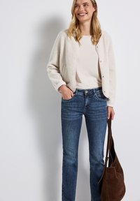 Cárdigan cremoso y texturizado sobre una camiseta clara, combinado con jeans de un azul medio. Sosteniendo un bolso de ante marrón. Diseño minimalista y materiales suaves.