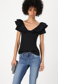 Top nero con scollatura a V e maniche a balze, abbinato a jeans azzurri chiari. Il tessuto appare liscio e aderente. Gli accessori includono una pochette.
