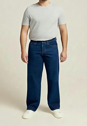 Muž v světle šedé košili s krátkými rukávy, tmavě modrých širokých džínách a bílých botách stojí před jednoduchým světlým pozadím.