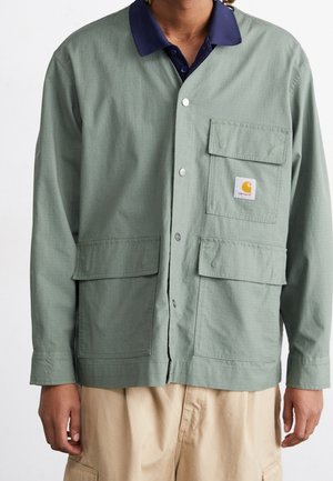 Persoon draagt een saliegroene blousejas met drie voorzakken en een witte logopatch, over een donkerblauw overhemd met kraag en beige broek.