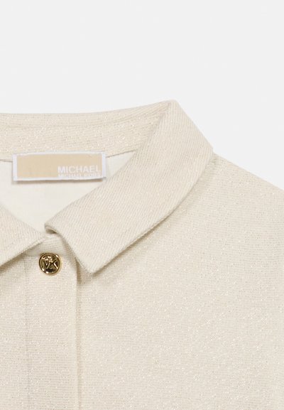 Μπεζ πουκάμισο από υφασμάτινη υφή με γιακά και χρυσό κουμπί χαραγμένο με "MK" και μια μπεζ ετικέτα Michael Kors μέσα στον λαιμό.