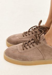 Zapatillas de ante marrón con punta redonda, suela de goma en contraste y cordones redondos. El diseño minimalista presenta acentos de costura.