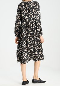 Robe noire à fleurs avec des manches longues, un col rond et une taille froncée. Conçue avec des fleurs crème et des feuilles vertes.
