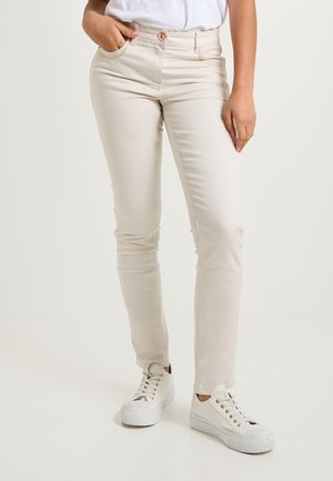 Jean skinny beige avec une fermeture à bouton, comportant des poches avant et un tissu en coton mélangé doux, associés à des baskets blanches.