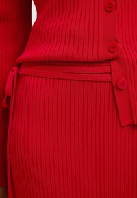 Top en tricot rouge à côtes associé à une jupe coordonnée. Présente des lignes verticales, cinq boutons sur le haut et un détail à nouer à la taille.