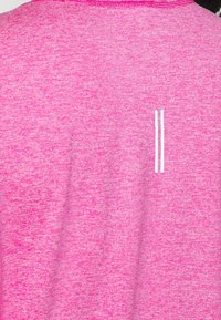 Vaaleanpunainen urheilupaita, joka on valmistettu teksturoidusta kankaasta ja jossa on meleerattu kuvio. Paidassa on valkoinen logo, jossa on kaksi pystysuoraa viivaa selässä.