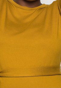 Un haut jaune moutarde avec une texture nervurée. Présente un col rond et une coupe à la taille avec une ceinture délicate.