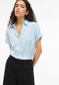 Ανοιχτό μπλε κοντομάνικο μπλουζάκι με κουμπιά στη λαιμόκοψη και γιακά, κατασκευασμένο από απαλό ύφασμα, σε συνδυασμό με μαύρο παντελόνι ραμμένο.