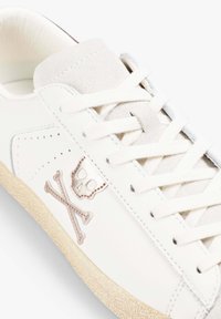 Sneaker in pelle bianca con lingua in suede, decorata con un design metallizzato di teschio e tibie incrociate, e suola beige testurizzata.