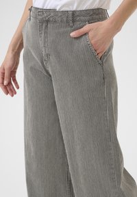 Henkilö, jolla on päällä pystysuoraraidalliset harmaat housut, oikea käsi taskussa ja vasen käsi rentona kyljessä yksinkertaista taustaa vasten.