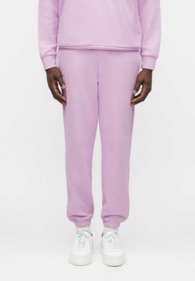 Osoba ubrana w jasnofioletowe spodnie dresowe i pasującą bluzę, stojąca z rękami swobodnie opuszczonymi wzdłuż ciała, widoczne białe trampki.