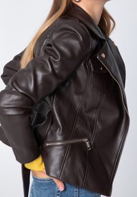 Ruskea nahkatakki, jossa on istuva malli, suuri kaulus, vetskarilla varustettu sivutasku ja näkyvät sauma-yksityiskohdat. Takissa on sileä, kiiltävä pinta.