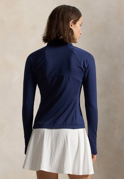 Γυναίκα με καστανά μαλλιά μέχρι τους ώμους, που φοράει μπλε σκούρη μπλούζα μακρυμάνικη και λευκή πλισέ φούστα με υφή, γυρισμένη προς τα πίσω μπροστά σε απλό φόντο.