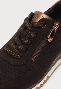 Gros plan d'une basket marron foncé avec des lacets aux accents cuivrés, du daim perforé, une fermeture éclair en cuivre et une semelle en caoutchouc texturée.