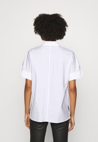 Persona con capelli ricci che indossa una camicia bianca a maniche corte e pantaloni neri, in piedi con le spalle alla camera su uno sfondo semplice.