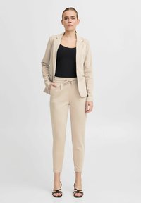 Beige blazer gecombineerd met een zwart tanktop en beige tapered broek met een trekkoord. Zwarte sandalen met bandjes maken de outfit compleet.