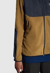 Fleecejack met een beige lichaam, marineblauwe accenten, een ritssluiting aan de voorkant en handzakken. Textuurdetail in de stof op de mouwen.