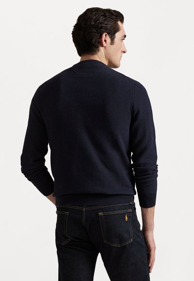 Suéter de punto azul marino con un escote redondo, puños acanalados y dobladillo, combinado con unos jeans de mezclilla oscuros que presentan detalles de costura amarilla.