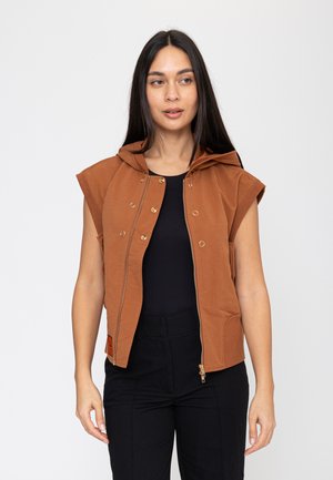 Vrouw met lang donker haar die een mouwloze bruine zip-up hoodie draagt over een zwart topje en zwarte broek, staand tegen een effen achtergrond.