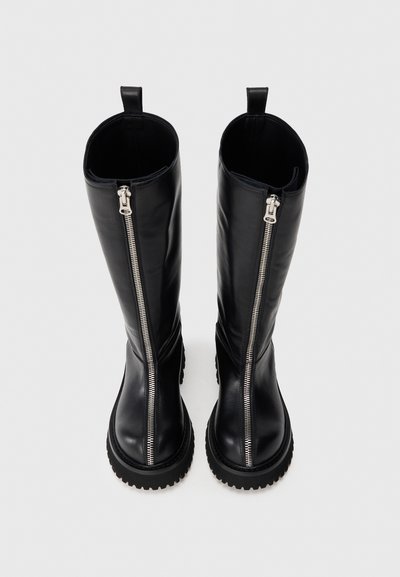 Bottes noires hauteur genoux avec des zips argentés à l'avant et des semelles épaisses à crampons, orientées vers l'avant sur un fond uni.