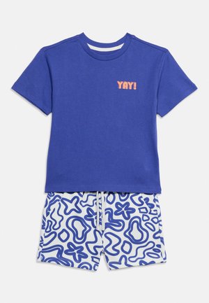 Blå kortærmet skjorte med teksten "YAY!" over hvide shorts med blå abstrakte mønstre og en hvid snørrebånds talje.